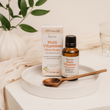 Laveen Multi Vitamine Olive Drops flesje met verpakking op een schaaltje, naast een houten lepel, met natuurlijke styling en groene bladeren op de achtergrond