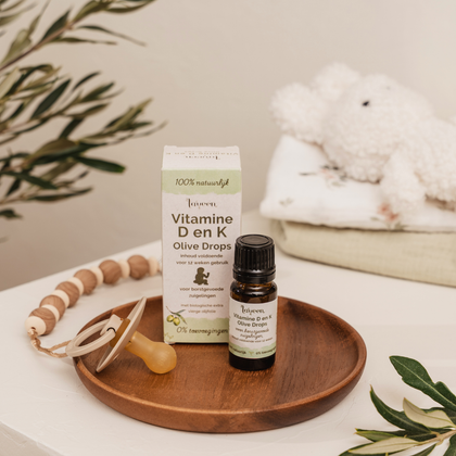 Flesje en verpakking van Laveen Vitamine D en K druppels voor zuigelingen op een houten schaaltje, met speen en babyaccessoires. Geschikt voor borstgevoede baby’s, op basis van biologische olijfolie en zonder toevoegingen.